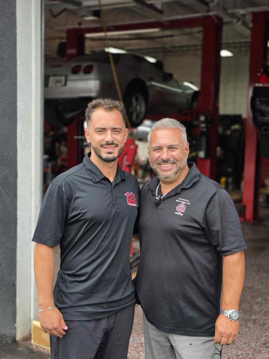 Two Automotive Solutions technicians under the shop sign with the bays open behind them